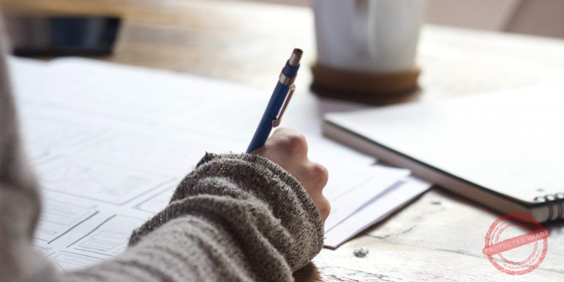 person writing on brown wooden table near white ceramic mug